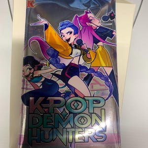 May include: A sealed K-Pop Demon Hunters trading card pack. The front features three anime-style female characters with microphones. The pack has a silver background with the title in large, iridescent letters. The top of the pack is blue.
