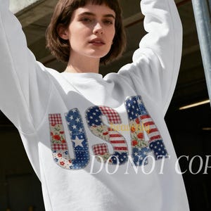 Op de afbeelding: Wit sweatshirt met het woord "USA" in een patchwork-ontwerp met de Amerikaanse vlag en bloempatronen. De sweatshirt heeft lange mouwen en een ronde hals. De tekst "DO NOT COPY" is zichtbaar op de sweatshirt.