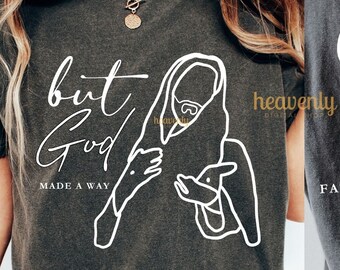 Op de afbeelding: Een donkergrijs T-shirt met een witte afbeelding van een persoon die een lam vasthoudt. De tekst "but God made a way" staat op de voorkant van het shirt. De achterkant van het shirt heeft de tekst "Jesus zei: "Ik ben de weg, de waarheid en het leven; niemand komt tot de Vader dan door mij." Johannes 14:6"