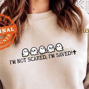 May include: A light beige sweatshirt with a line of cute cartoon ghosts and the text "I'M NOT SCARED, I'M SAVED!+"