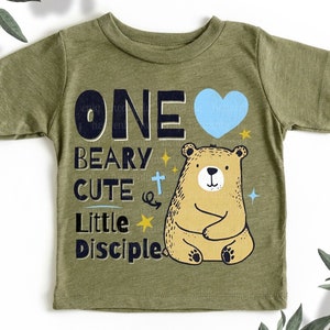 May include: A green short-sleeved t-shirt with a bear and the text "ONE BEARY CUTE LITTLE DISCIPLE" in black and blue. A blue heart is next to the word "ONE".