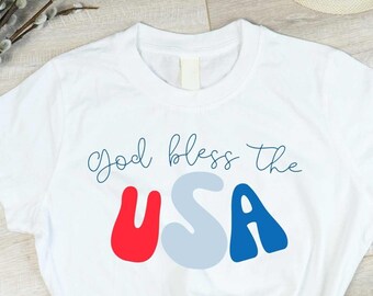 May include: White t-shirt with the text "God bless the USA" in blue, red, and gray lettering. The letters are stylized and rounded.