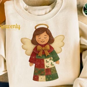 Puede incluir: Sudadera color crema con un diseño de ángel navideño. El ángel tiene cabello castaño, un halo dorado y alas beige. El vestido del ángel es de retazos con patrones rojos, verdes y dorados. El texto "Heavenly" está impreso en la parte superior izquierda.