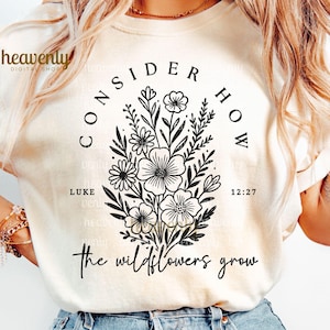 May include: Cream-colored t-shirt featuring a black floral design with the text "CONSIDER HOW" and "the wildflowers grow." The design includes the text "LUKE 12:27." The shirt has short sleeves and a relaxed fit.