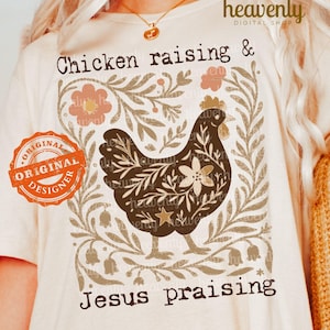 Può includere: T-shirt color crema con illustrazione di gallina marrone e dettagli floreali. Il testo "Chicken raising & Jesus praising" è visualizzato in un carattere decorativo. È visibile un timbro "Original Designer".