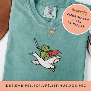 May include: A sage green t-shirt featuring an embroidered graphic of a green frog wearing a red cape and holding a sword while riding a white goose. The shirt has a Comfort Colors label and a circular overlay with the text Heavenly Embroidery Files 4 Sizes.