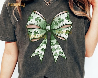 May include: A dark gray t-shirt with a green shamrock bow design. The bow is made of a green fabric with a white outline and is tied in a knot. The shamrocks are scattered throughout the bow.