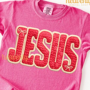 May include: A pink t-shirt with the word "JESUS" in a red knit-style font with a cream-colored outline. The shirt has a crew neck and short sleeves. The brand name "Comfort Colors" is visible on the tag.