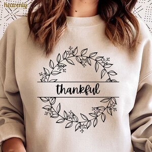 May include: A beige sweatshirt with the word "thankful" in black script, framed by a black floral wreath design. The sweatshirt is a neutral color, suitable for fall or everyday wear. The design is simple and elegant.