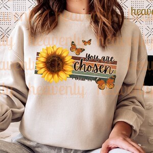 May include: A beige sweatshirt with a vintage-style graphic design featuring a sunflower, butterflies, and the text "You are Chosen" with the scripture reference "1 Thess. 1:4".