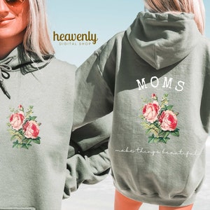 May include: Sage green hoodie with a floral design of pink roses and green leaves. The front of the hoodie features the floral design, while the back displays the word "MOMS" above the floral design and the phrase "make things beautiful."