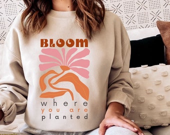 Puede incluir: Sudadera beige con las palabras "BLOOM where you are planted" en una fuente retro. El gráfico presenta una flor rosa y formas abstractas naranjas. La sudadera es de color neutro y tiene cuello redondo.