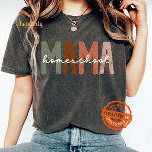 Puede incluir: Una camiseta gris oscura con el texto "MAMA homeschool" en letras coloridas y desgastadas. Las letras son en tonos de verde, naranja, rosa y rojo. La camiseta tiene un aspecto desgastado y la lleva puesta una persona con el pelo largo y casta&ntilde;o. La imagen tambi&eacute;n incluye una peque&ntilde;a pegatina circular naranja con el texto "ORIGINAL DESIGNER" en blanco.