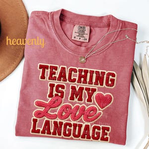 K&ouml;nnte beinhalten: Ein staubrosafarbenes T-Shirt mit dem Schriftzug "TEACHING IS MY LOVE LANGUAGE" in roter und cremefarbener Schrift. Eine silberne Halskette mit Anh&auml;nger liegt auf dem Shirt. Das Shirt ist gefaltet und auf einer wei&szlig;en Oberfl&auml;che ausgestellt.