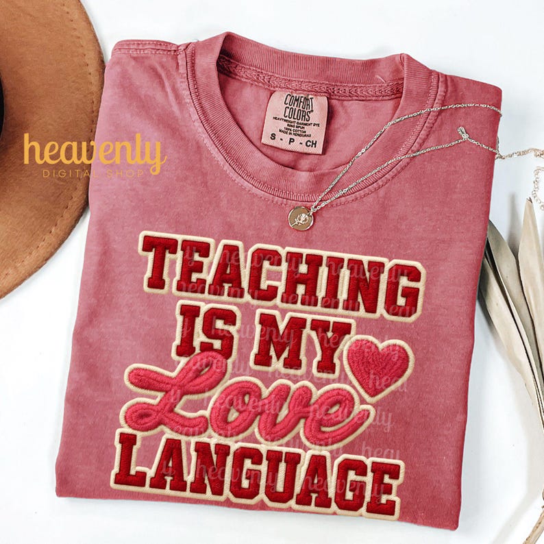 K&ouml;nnte beinhalten: Ein staubrosafarbenes T-Shirt mit dem Schriftzug "TEACHING IS MY LOVE LANGUAGE" in roter und cremefarbener Schrift. Eine silberne Halskette mit Anh&auml;nger liegt auf dem Shirt. Das Shirt ist gefaltet und auf einer wei&szlig;en Oberfl&auml;che ausgestellt.