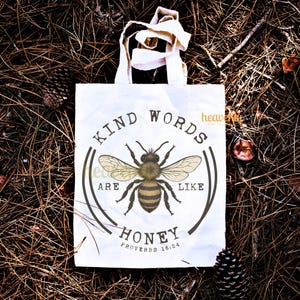 May include: White tote bag with a circular design featuring a detailed bee illustration. The text reads "KIND WORDS ARE LIKE HONEY" with "PROVERBS 16:24" below. The bag has cream-colored handles.