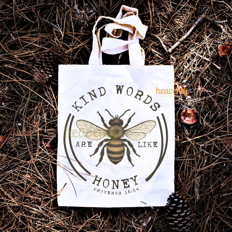 May include: White tote bag with a circular design featuring a detailed bee illustration. The text reads "KIND WORDS ARE LIKE HONEY" with "PROVERBS 16:24" below. The bag has cream-colored handles.