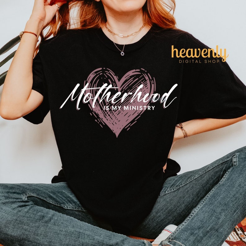 Op de afbeelding: Zwart T-shirt met een roze hartgrafiek en de tekst "Motherhood is my ministry".