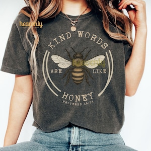 May include: Dark gray t-shirt with a circular graphic featuring a bee illustration. The text reads "Kind Words Are Like Honey" with "Proverbs 16:24" below. The shirt has short sleeves and a crew neck.