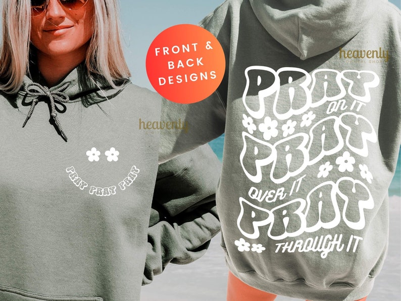 May include: A sage green hoodie with white text and flower graphics. The front features a smiley face design with the words "PRAY PRAY PRAY". The back displays "PRAY on it, PRAY over it, PRAY through it".