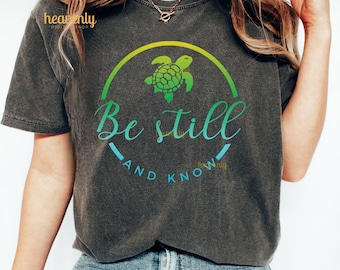 Puede incluir: Una camiseta gris oscura con un gráfico de tortuga marina verde y el texto "Be still and know" en un círculo.