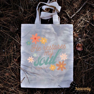 Puede incluir: Una bolsa de lona blanca con un dise&ntilde;o floral y el texto "He restores my soul" con la referencia b&iacute;blica "Salmo 23:3".