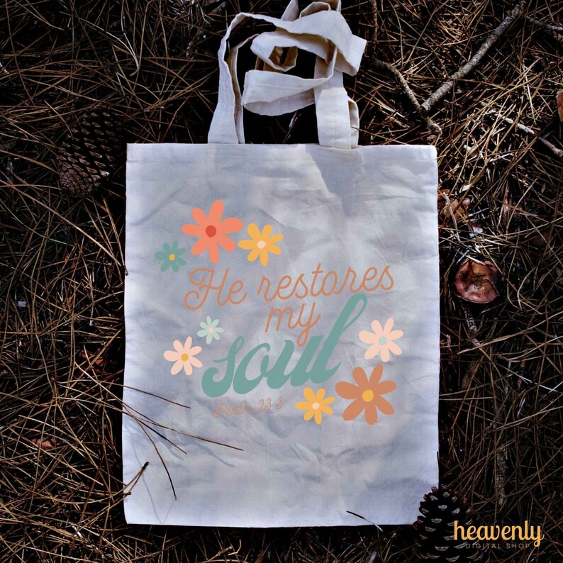 Puede incluir: Una bolsa de lona blanca con un dise&ntilde;o floral y el texto "He restores my soul" con la referencia b&iacute;blica "Salmo 23:3".