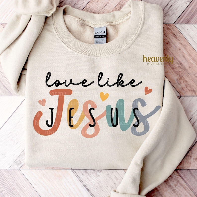 May include: A cream-colored sweatshirt with the words "love like Jesus" in black script. The word "Jesus" is in colorful letters with small heart accents. The brand tag is visible on the upper center of the sweatshirt.