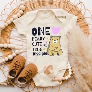 May include: A cream-colored baby onesie with a graphic of a bear and the text "ONE BEARY CUTE LITTLE DISCIPLE" in black. The bear is sitting down and has a pink heart above it.