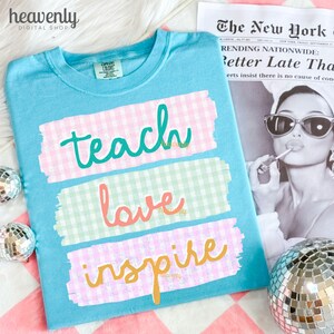 May include: A light blue t-shirt with the words "teach, love, inspire" in a colorful, playful font. The words are layered on top of pastel gingham patterns. The shirt is displayed with a vintage newspaper and disco balls.