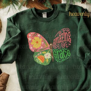 Puede incluir: Una sudadera verde oscuro con un dise&ntilde;o de mariposa colorida. La mariposa tiene un patr&oacute;n floral rosa, amarillo y verde y las palabras "Amazing Grace" escritas en sus alas.