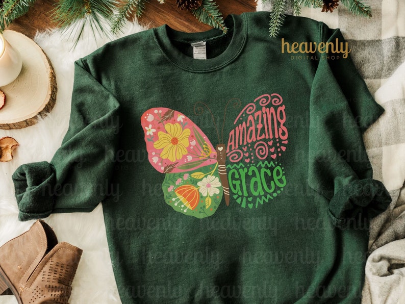 Puede incluir: Una sudadera verde oscuro con un dise&ntilde;o de mariposa colorida. La mariposa tiene un patr&oacute;n floral rosa, amarillo y verde y las palabras "Amazing Grace" escritas en sus alas.