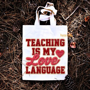 K&ouml;nnte beinhalten: Eine wei&szlig;e Tragetasche mit dem Text "TEACHING IS MY LOVE LANGUAGE" in roter und cremefarbener Schrift. Die Tasche hat wei&szlig;e Griffe und liegt auf einem Bett aus Kiefernadeln und Tannenzapfen.