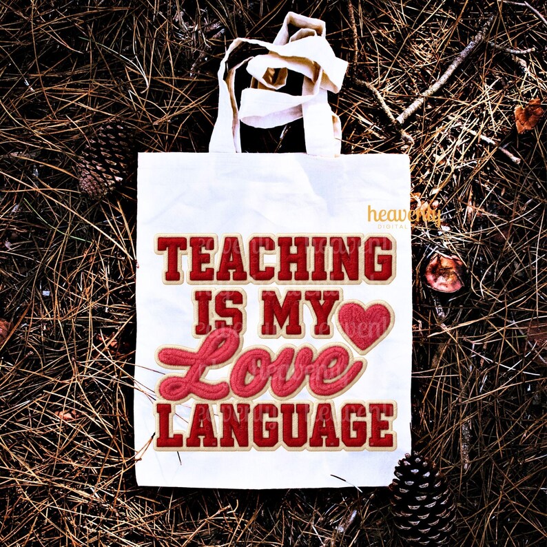 K&ouml;nnte beinhalten: Eine wei&szlig;e Tragetasche mit dem Text "TEACHING IS MY LOVE LANGUAGE" in roter und cremefarbener Schrift. Die Tasche hat wei&szlig;e Griffe und liegt auf einem Bett aus Kiefernadeln und Tannenzapfen.