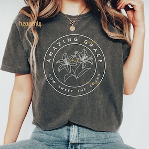 Pu&ograve; includere: T-shirt grigio scuro con la scritta "AMAZING GRACE" in un design circolare, con due illustrazioni di fiori di giglio bianchi. &Egrave; inclusa anche la frase "HOW SWEET THE SOUND". La maglietta &egrave; realizzata in un materiale morbido.