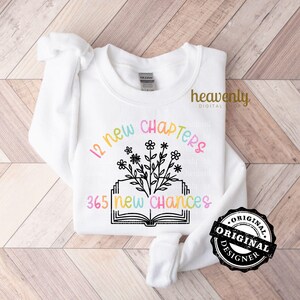 May include: A white sweatshirt with a black graphic design of an open book with flowers and the text "12 new chapters 365 new chances".