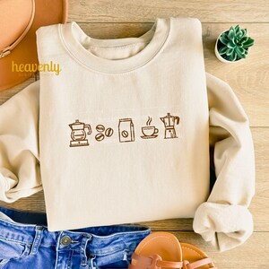 May include: A beige sweatshirt with a brown graphic of coffee beans, a coffee bag, a cup of coffee, and a coffee pot.