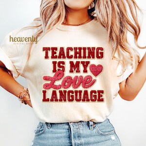 K&ouml;nnte beinhalten: Cremefarbenes T-Shirt mit dem Text "TEACHING IS MY LOVE LANGUAGE" in roter und rosa Schrift. Das Shirt hat eine Herzgrafik und wird mit einer blauen Jeans getragen.