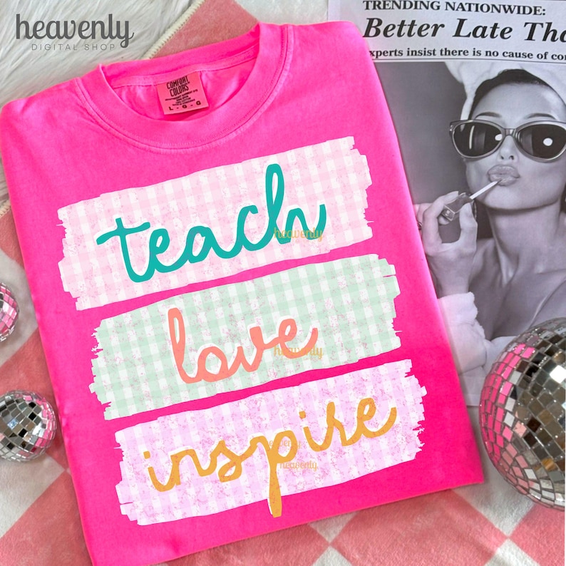 May include: A bright pink t-shirt with the words "teach, love, inspire" in teal, coral, and yellow script. The words are on white rectangular brushstrokes with a gingham pattern. The shirt has a Comfort Colors tag.