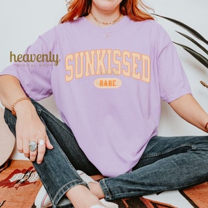 May include: A lavender t-shirt with the text "SUNKISSED the BABE" in orange and yellow lettering.
