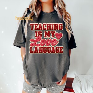 K&ouml;nnte beinhalten: Dunkelgraues T-Shirt mit dem Text "TEACHING IS MY LOVE LANGUAGE" in roter und cremefarbener Schrift. Das Shirt hat kurze &Auml;rmel und eine lockere Passform. Das Design beinhaltet ein Herzsymbol.