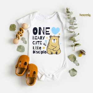 May include: White baby onesie with a blue heart, a brown bear, and the text "ONE BEARY CUTE LITTLE DISCIPLE".