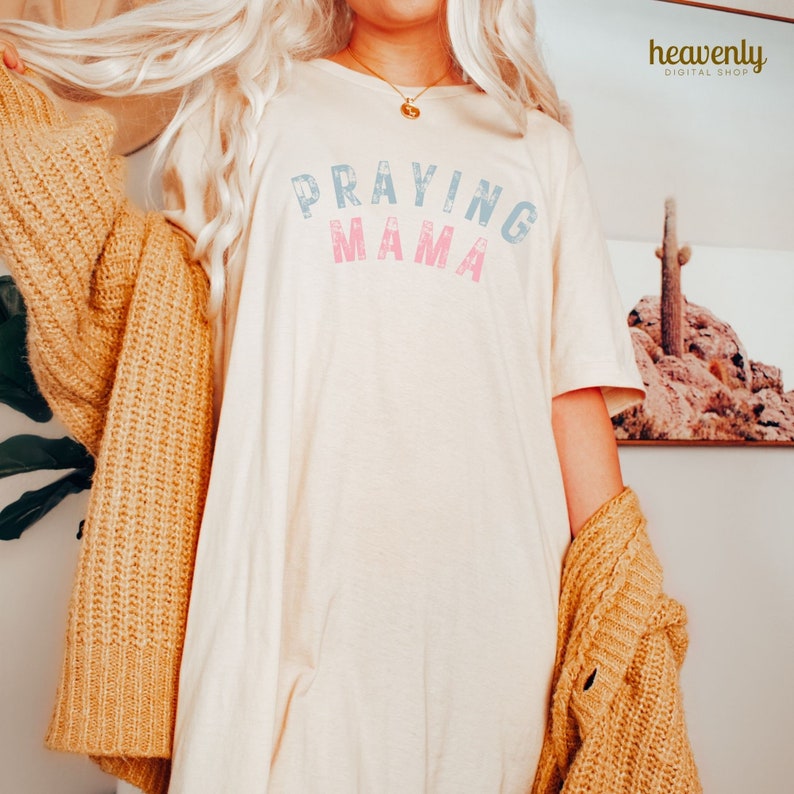 May include: A white t-shirt with the text "Praying Mama" in blue and pink lettering. The shirt is being worn by a person with blonde hair and a brown cardigan.