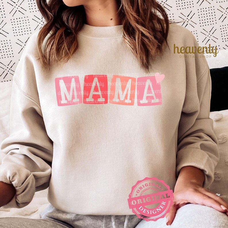 May include: A light beige sweatshirt with the word "MAMA" printed in pink, red, and orange block letters with a small pink heart to the right of the word.