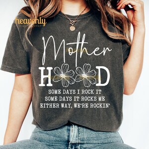 May include: A dark gray t-shirt with the text "Mother Hood" in white with three white flowers in the middle of the word "Hood". The text "Some days I rock it, some days it rocks me, either way, we're rockin'" is printed below the main text.