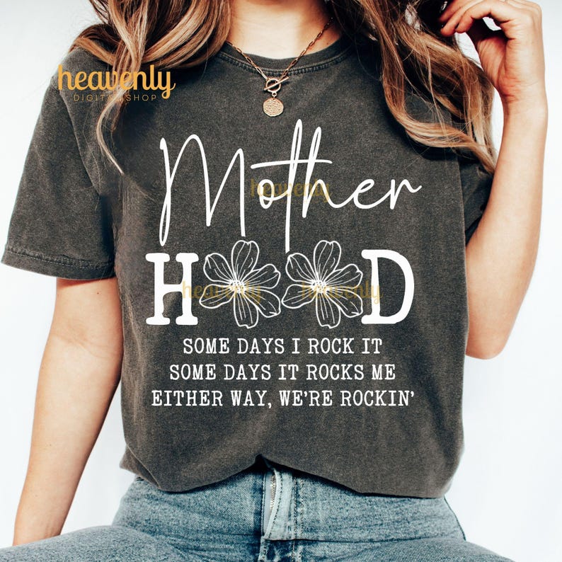 May include: A dark gray t-shirt with the text "Mother Hood" in white with three white flowers in the middle of the word "Hood". The text "Some days I rock it, some days it rocks me, either way, we're rockin'" is printed below the main text.