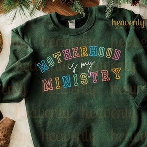 Pu&ograve; includere: Una felpa verde scuro con il testo "MOTHERHOOD is my MINISTRY" in colori arcobaleno. Il testo &egrave; contornato di nero e ha un aspetto retr&ograve;, vintage.