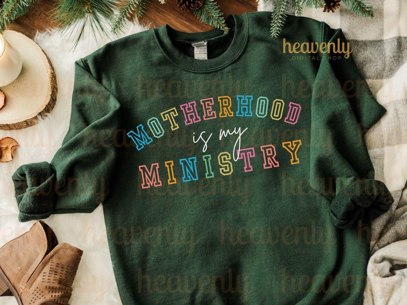 Pu&ograve; includere: Una felpa verde scuro con il testo "MOTHERHOOD is my MINISTRY" in colori arcobaleno. Il testo &egrave; contornato di nero e ha un aspetto retr&ograve;, vintage.