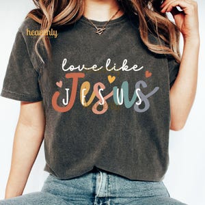 May include: Dark gray t-shirt with the words "love like Jesus" in white script above the word "Jesus" in colorful block letters. Small heart graphics are scattered around the text. The shirt is a casual style.