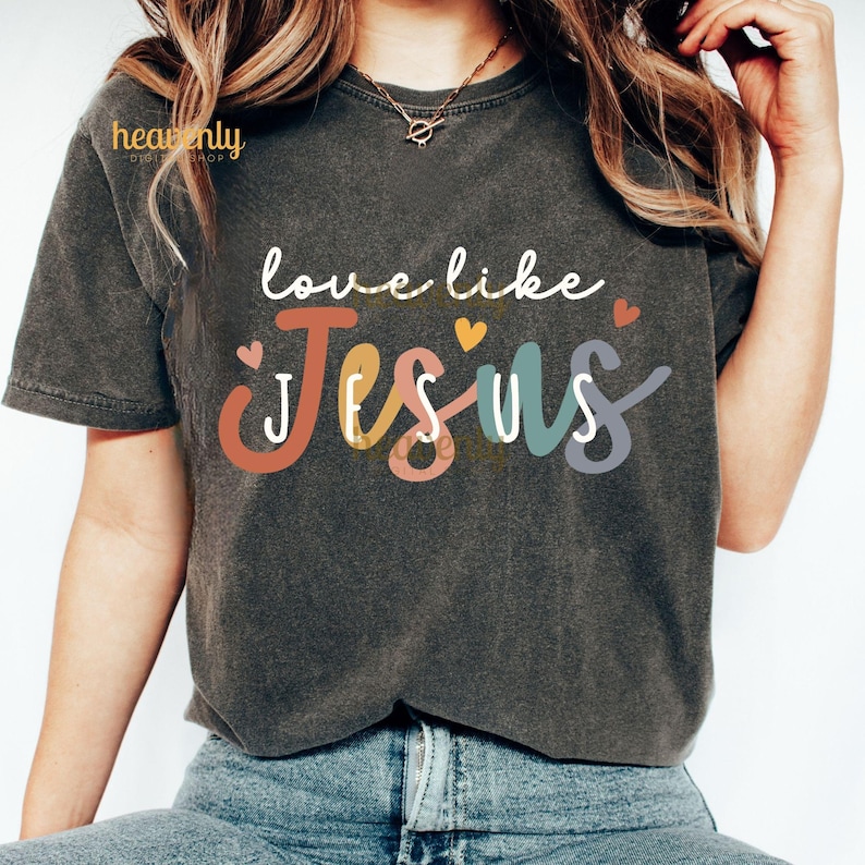 May include: Dark gray t-shirt with the words "love like Jesus" in white script above the word "Jesus" in colorful block letters. Small heart graphics are scattered around the text. The shirt is a casual style.